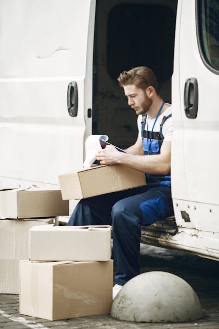 A man wearing a blue and white striped shirt and dark work trousers is seated at the open side door of a white moving van, surrounded by several cardboard boxes of varying sizes, some stacked on the ground and others held in his hands. The boxes are plain brown, with some partially covered in protective packing materials like plastic wrap or paper. The man is holding a clipboard or documents, possibly reviewing inventory or moving instructions, in preparation for a home relocation. The van's interior is visible behind him, showing a dark cargo space ready for packing or loading furniture and household items. The scene is set outdoors on a paved driveway or street, with a concrete barrier or bollard nearby, indicating a typical moving environment for furniture transport and packing processes, and reflects a professional move managed by [COMPANY_NAME].