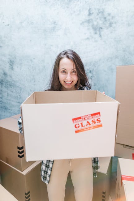 A woman with long dark hair smiling at the camera while holding an open, empty cardboard box with a red and white 'Fragile Glass' sticker on the front, during a home relocation. She is standing inside a room with a textured light blue wall, surrounded by several packed cardboard boxes of various sizes, some sealed with tape, placed on the floor and nearby surfaces. The scene depicts the process of packing and moving, with visible materials such as cardboard, packing tape, and packaging labels, and equipment like the open box being prepared for transport. Man and Van Eastcote's removal services involve careful packing, loading, and handling of items like glassware, illustrated here by the labeled box, as part of a furniture transport or home moving operation.