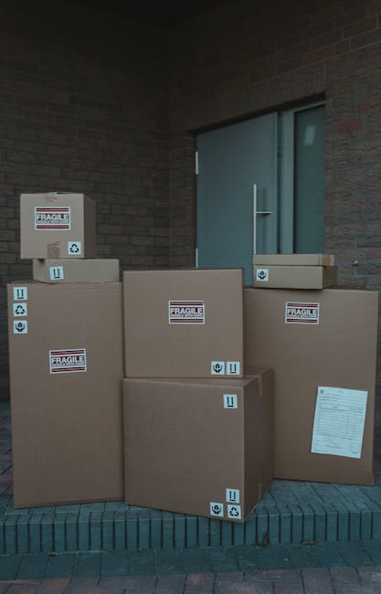 A man wearing a blue and white striped shirt and dark work trousers is seated at the open side door of a white moving van, surrounded by several cardboard boxes of varying sizes, some stacked on the ground and others held in his hands. The boxes are plain brown, with some partially covered in protective packing materials like plastic wrap or paper. The man is holding a clipboard or documents, possibly reviewing inventory or moving instructions, in preparation for a home relocation. The van's interior is visible behind him, showing a dark cargo space ready for packing or loading furniture and household items. The scene is set outdoors on a paved driveway or street, with a concrete barrier or bollard nearby, indicating a typical moving environment for furniture transport and packing processes, and reflects a professional move managed by [COMPANY_NAME].