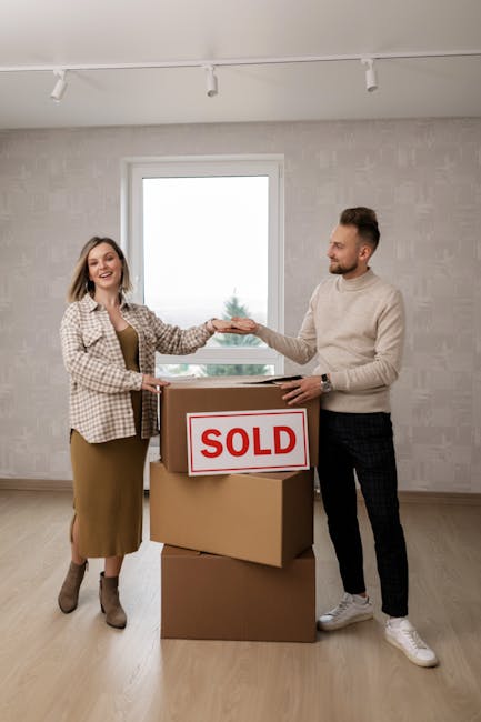 A woman and a man are standing inside a well-lit room, engaged in a house relocation process. The woman, dressed in a beige skirt and checkered blazer, is smiling and holding a small package or document, handing it over to the man. The man, wearing a light-colored sweater and dark checkered trousers, is reaching out to accept the item, with a gentle smile. They are positioned next to a large cardboard box marked with a red 'SOLD' sign, which is placed on top of another cardboard box, indicating a recent property transaction or move. The room has a large window in the background, letting in natural light, and plain walls with smooth flooring. This scene captures the process of furniture transport, packing, or moving logistics, typical of house removals, with the involvement of professional services like those offered by Man and Van Eastcote. No specific moving equipment is visible, but the context suggests an organized handover during a home relocation.