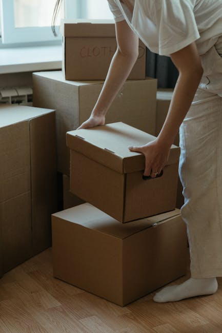 A woman and a man are standing inside a well-lit room, engaged in a house relocation process. The woman, dressed in a beige skirt and checkered blazer, is smiling and holding a small package or document, handing it over to the man. The man, wearing a light-colored sweater and dark checkered trousers, is reaching out to accept the item, with a gentle smile. They are positioned next to a large cardboard box marked with a red 'SOLD' sign, which is placed on top of another cardboard box, indicating a recent property transaction or move. The room has a large window in the background, letting in natural light, and plain walls with smooth flooring. This scene captures the process of furniture transport, packing, or moving logistics, typical of house removals, with the involvement of professional services like those offered by Man and Van Eastcote. No specific moving equipment is visible, but the context suggests an organized handover during a home relocation.