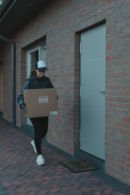 A woman wearing a baseball cap, glasses, a dark jacket, and black leggings is carrying a medium-sized cardboard box labeled 'fragile' in black text with handling instructions, as she walks along a brick-paved pathway outside a residential building. She is approaching a white door set into a red brick wall, with a black doormat in front of it. The scene suggests an outdoor home relocation or moving process orchestrated by Man and Van Eastcote, with the woman preparing to load or unload furniture or boxes during packing and moving activities, under natural lighting conditions.