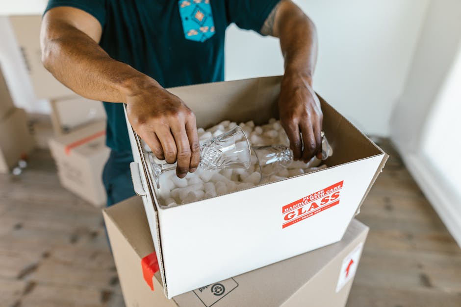 A person wearing a blue shirt with a patterned tie is seen packing a fragile glass object into a cardboard box lined with white packing peanuts. The box is labelled with a red sticker that reads 'HANDLE WITH CARE GLASS.' The individual is placing the glass carefully inside, with their hands visible gripping the item and stabilizing it during the packing process. The scene takes place indoors, with a wooden floor and a white wall in the background. Partially visible in the foreground is another box under the packing activity, and cardboard boxes are stacked nearby, consistent with home relocation or furniture transport activities. The lighting is natural, indicating daylight, and the focus is on the careful packing of fragile items during a moving or packing and moving process, typical of professional removals by Man and Van Eastcote.