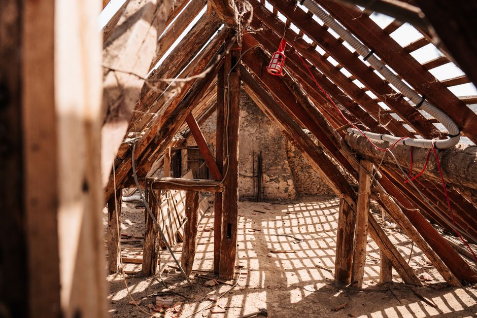 Inside an unfinished or abandoned attic space, showing exposed wooden roof trusses and beams, some of which are supported by vertical wooden posts. The roof has gaps allowing natural light to enter, i