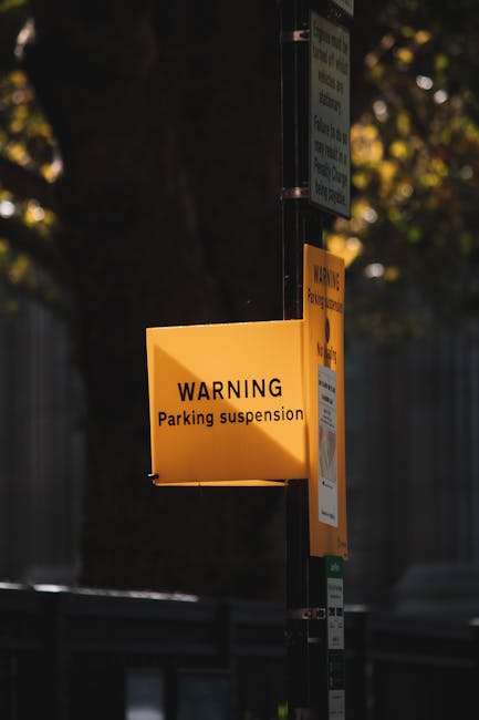 A yellow warning sign attached to a black metal pole displays the message 'WARNING Parking suspension' in black text, indicating temporary parking restrictions in place around the area. The sign is positioned outdoors, with a background featuring a blurred tree with green and yellow leaves and a grey fence or wall, suggesting an urban or residential environment. Above the warning sign, additional smaller notices are visible, possibly detailing specific parking restriction times or dates, attached to the same pole. The scene appears to be captured during daylight hours with natural lighting, and the sign is in focus, providing clear visibility of the message. This image relates to parking management, which is relevant in the context of house removals, relocation services, and moving logistics, as indicated by the webpage title 'Eastcote parking restrictions Hillingdon Council removals, EASTCOTE' and the company's focus, Man and Van Eastcote.