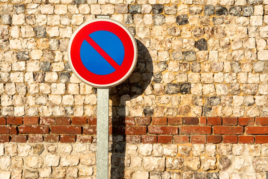 A circular no parking traffic sign with a blue background and a red border, mounted on a metal pole, positioned in front of a textured brick wall composed of light and dark beige stones with a horizontal red brick line. Behind the sign, there is a paved area with several cardboard boxes, some wrapped in plastic and others open, containing items like small furniture and household goods. A man wearing casual clothing is seen lifting and carrying a cardboard box towards a white van parked nearby, which is partially visible with its side door open. The scene appears to depict a home relocation or furniture transport process outside a residential property, with the focus on loading items into the vehicle, supported by the presence of packing materials, furniture, and transport equipment such as trolleys or straps. The lighting suggests daytime with clear weather, and the overall environment is an outdoor area adjacent to a house or building, aligning with house removals or moving services provided by [COMPANY_NAME].