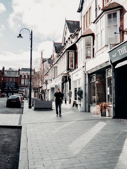 A man dressed in dark clothing is walking on a paved sidewalk in front of a row of traditional white-fronted shopfronts with large windows, some adorned with decorative plants. To the left, a streetlamp with a curved arm extends over the sidewalk, and parked cars are visible along the curb. The shops are part of a retail street, with one shop having a black awning displaying white lettering, and another with a white door and window displays. The scene is captured during daylight with clear weather, and the urban environment suggests a typical shopping and commercial area in Eastcote. The presence of this setting in the context of house removals and furniture transport underscores the first stage of a home relocation involving packing and moving items through such retail streets, with a focus on loading and logistics handled by [COMPANY_NAME].