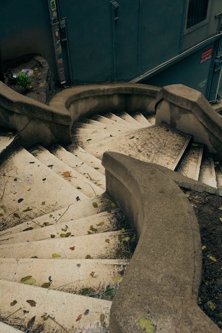 A curved concrete staircase outside a building, leading downward with visible wear and some fallen leaves scattered on the steps. The staircase has a rounded handrail on one side and a concrete wall on the other. Behind the stairs, there are blue metal utility cabinets and a part of a gray building. The image appears to be taken during daylight, with natural light illuminating the area. The setting suggests an entry or exit point of a property where furniture or boxes could be moved during a home relocation, with the stairs potentially used to carry items between street level and the entrance. The overall scene captures a typical residential urban environment, with elements relevant to the process of house removals handled by services such as Man and Van Eastcote.