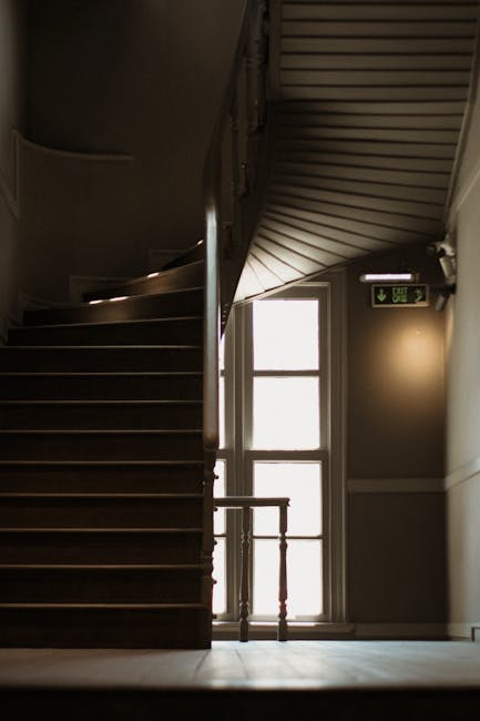 A narrow staircase inside a property with dark wooden steps and a curved handrail ascending from the ground floor. To the right, there is a large window allowing natural light to illuminate the area, next to a closed white door with multiple glass panes. The staircase is adjacent to a wall painted in a neutral tone, and an emergency exit sign with a green running figure and an arrow is mounted high on the wall near the ceiling. The scene captures the interior environment where a home relocation or furniture transport process might be taking place, with no furniture or moving equipment visible in this specific view. The detailed description highlights the spatial arrangement and lighting conditions typical of residential hallways involved in packing and moving activities, as handled by professional removals services such as Man and Van Eastcote.