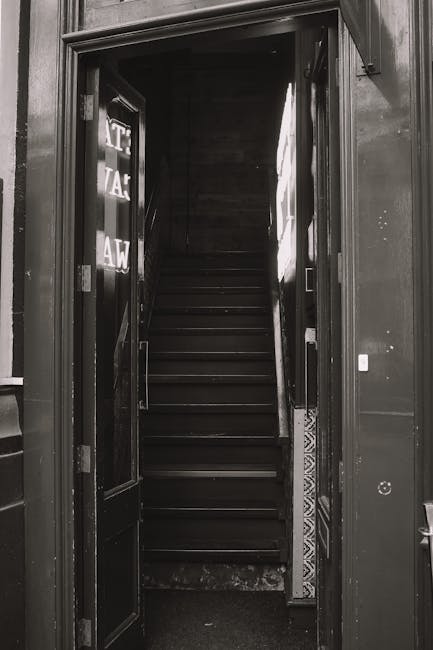 A curved concrete staircase outside a building, leading downward with visible wear and some fallen leaves scattered on the steps. The staircase has a rounded handrail on one side and a concrete wall on the other. Behind the stairs, there are blue metal utility cabinets and a part of a gray building. The image appears to be taken during daylight, with natural light illuminating the area. The setting suggests an entry or exit point of a property where furniture or boxes could be moved during a home relocation, with the stairs potentially used to carry items between street level and the entrance. The overall scene captures a typical residential urban environment, with elements relevant to the process of house removals handled by services such as Man and Van Eastcote.
