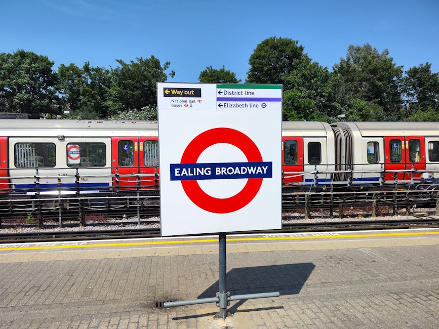 A large white sign with the London Underground roundel and the text 'Ealing Broadway' in blue is situated on a paved platform at a train station. Behind the sign, a modern electro-mechanical train with red, white, and blue livery is stationary on the tracks, with visible windows and doors. To the left, a yellow and black directional arrow signs towards the 'Way out,' with additional signs indicating the district and Elizabeth lines, as well as connections to national rail and bus services. The platform is adjacent to railway tracks with safety rails separating the platform from the trains. In the background, lush green trees under a clear blue sky suggest a sunny day. This scene is consistent with a London tube station, captured during a typical home relocation or furniture transport process that involves coordinating station access and car or van transit managed by companies like Man and Van Eastcote.