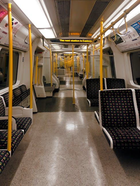 The image depicts the interior of a modern train carriage with a clean, well-lit environment featuring rows of fabric-covered seats with a geometric multicolour pattern. Yellow vertical poles and overhead handrails are positioned along the aisle for passenger stability, with some poles extending to the ceiling. The carriage has large rectangular windows on both sides, allowing natural light to illuminate the space. Above the aisle, an electronic display shows the message 'The next station is Eastcote,' indicating the approaching stop. The flooring is a neutral, textured material suitable for high foot traffic. Although there are no visible luggage or packaging materials, the scene emphasizes the spaciousness and efficient layout of the train interior. This setting aligns with the context of relocation services, with the train environment suggesting transport or travel related to home moves or furniture transport, which Man and Van Eastcote often facilitates during house removals near Eastcote Tube Station, especially considering delays or logistics challenges.