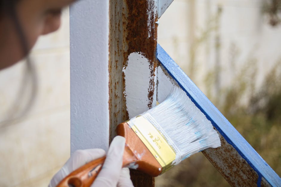 A person wearing white gloves is applying white paint with a broad brush to the corner of a wooden doorframe, which is partially rusted and weathered. The doorframe is being prepared for house repairs or refreshment, with blue painter's tape applied along the edge to create a clean division between the painted surface and the unpainted wood. The background shows an out-of-focus outdoor setting with blurred trees and natural light, indicating a daytime project. This scene is part of a home improvement or renovation process, which might be relevant to house removals or property preparations handled by Man and Van Eastcote, especially during packing or moving preparations involving home upgrades.