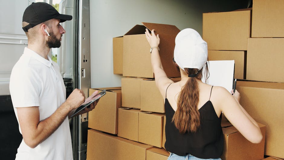 A man with curly hair and wearing a blue t-shirt and dark trousers sits on a wooden floor inside a room with large, arched windows that allow natural daylight to illuminate the space. Several cardboard boxes, some sealed with red packing tape, are stacked nearby, suggesting recent packing or ongoing packing activities as part of a home relocation process. The boxes are positioned against the wall behind the man, who appears to be resting during a furniture transport session. To the right, a tall potted plant with broad green leaves is placed next to the window, adding a touch of interior decor. Outside the windows, a street view with parked cars and multi-storey buildings is visible, indicating an urban environment. The natural light highlights the wooden flooring and creates a calm atmosphere typical of packing and moving procedures. This scene exemplifies a staged moment during professional removals, such as those offered by Man and Van Eastcote, supporting the process of packing, loading, or planning a house move.