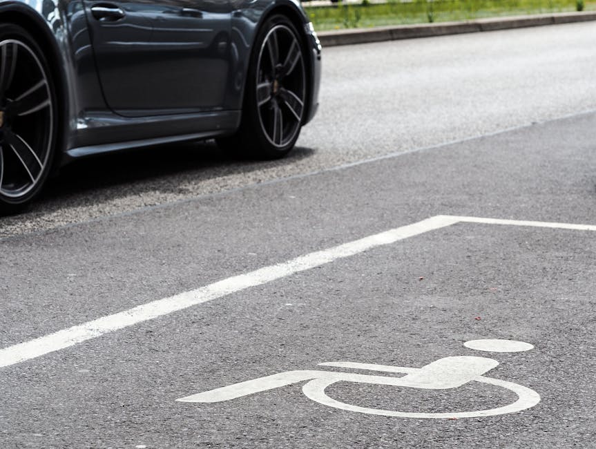 Close-up view of a black sports car parked on a paved street with a dedicated parking space marked by white lines, including a wheelchair symbol painted on the asphalt. The car's front wheel and part of the vehicle's bodywork are visible, showing sleek design and alloy wheels. The parking area is adjacent to a property with an access point, relevant to home relocation and furniture transport. The scene highlights the importance of designated parking zones for moving vehicles and equipment during house removals, with the street's textured asphalt surface and natural lighting providing clarity for accessibility and logistical planning. As part of the moving process, flexible parking arrangements and clear markings assist movers from [COMPANY_NAME] during packing, loading, and furniture transport in the Eastcote area, ensuring efficient and safe removal services.