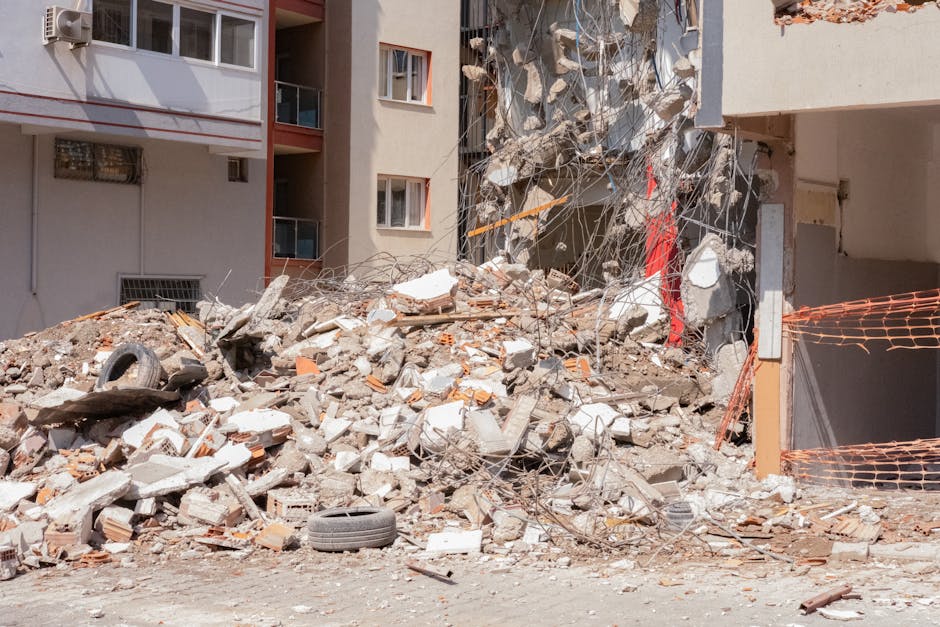 Photograph of a building demolition site showing a partially collapsed multi-storey structure with exposed concrete and steel rebar. The debris includes broken bricks, concrete fragments, twisted metal wires, and scattered building materials. An orange plastic safety fence is visible on the right side, with the ground covered in rubble and large chunks of concrete. There are a few discarded vehicle tires among the debris, and nearby, part of the building’s exterior wall still stands with several windows intact. The scene is outdoors during daytime with natural light illuminating the dusty environment, illustrating a typical urban demolition process that may require proper removal permits. Man and Van Eastcote occasionally handles logistical aspects of such site clearances as part of their house removals and relocation services, ensuring safe and efficient transport of household items once the site is ready for building work or refurbishment.