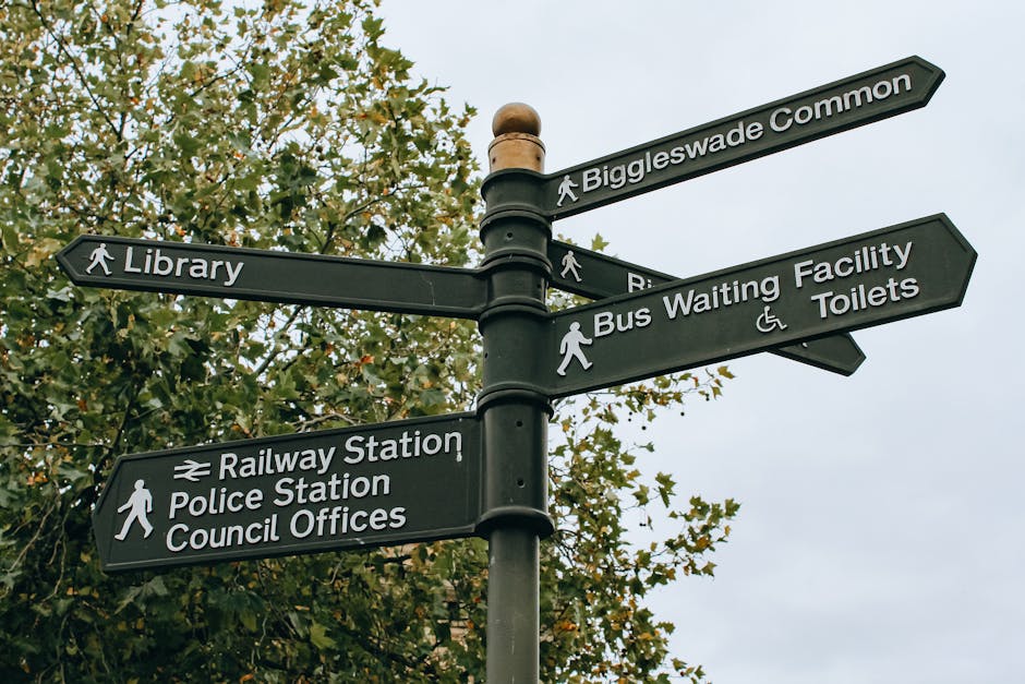 A black multi-directional signpost situated outdoors against a background of leafy trees and an overcast sky, with white lettering and icons indicating directions to local amenities such as Biggleswade Common, Library, Railway Station, Police Station, Council Offices, Bus Waiting Facility, and Toilets, including accessibility symbols for wheelchair users and pedestrians. The signpost is mounted on a metal pole with several directional arms pointing in different directions, each labeled clearly for ease of navigation. This scene relates to community infrastructure and local navigation in Eastcote, relevant to house removals and relocation services provided by Man and Van Eastcote, especially when planning moves that involve accessing municipal facilities or public transport options during a home relocation process.
